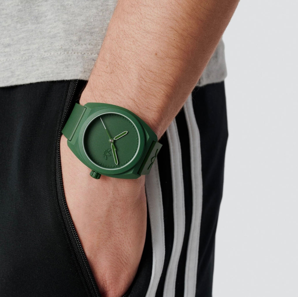 Adidas AOST24053 green watch worn on a wrist beside dark striped trousers.