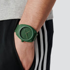 Adidas AOST24053 green watch worn on a wrist beside dark striped trousers.