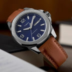 Timex Mens Watch Essex Avenue TW2W92800, blue dial, silver case, brown leather strap, resting on a brown surface at a slight angle.